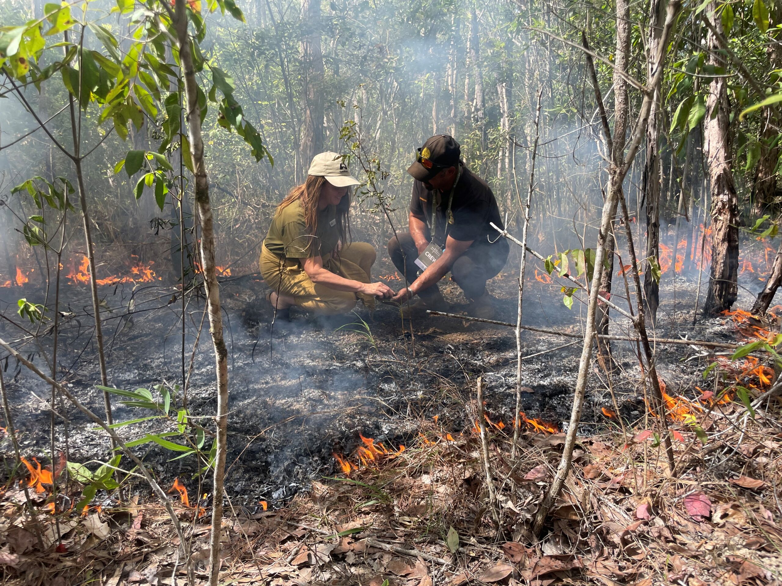 2 participants crouching and looking down at burnt land with a low and slow burn fire surrounding them. Small trees around.